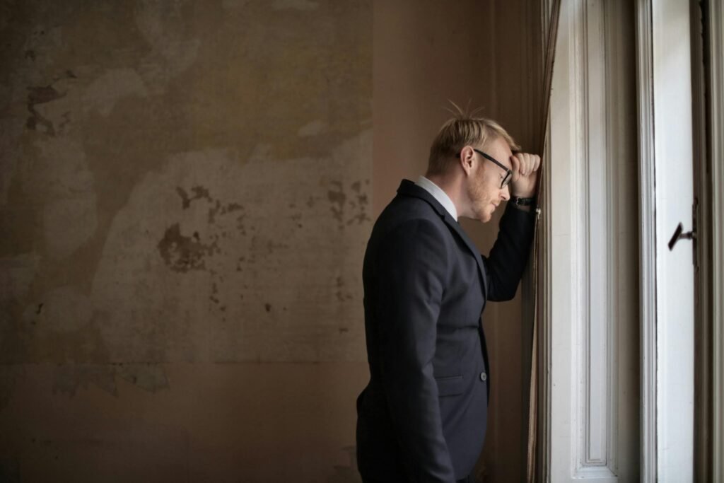 Side view of hopeless young businessman in classy outfit standing near window in shabby house and leaning on hand near wall while realizing financial losses
